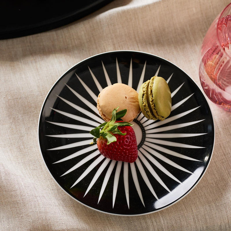 A black and white geometric patterned Marrakech Small Plates by Caskata Artisanal Home holds a strawberry and two macarons, one beige and one green, placed on a neutral-colored fabric.