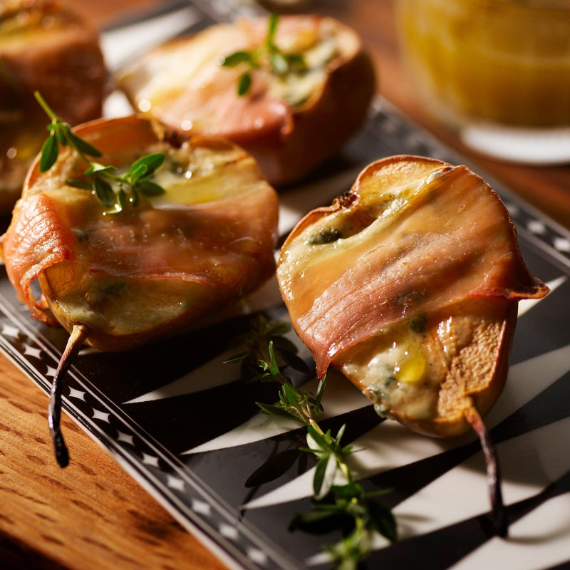 Pear halves grilled and wrapped in crispy bacon with melted cheese, garnished with herbs, presented elegantly on Caskata's Marrakech Petite Trays, Set of 2.