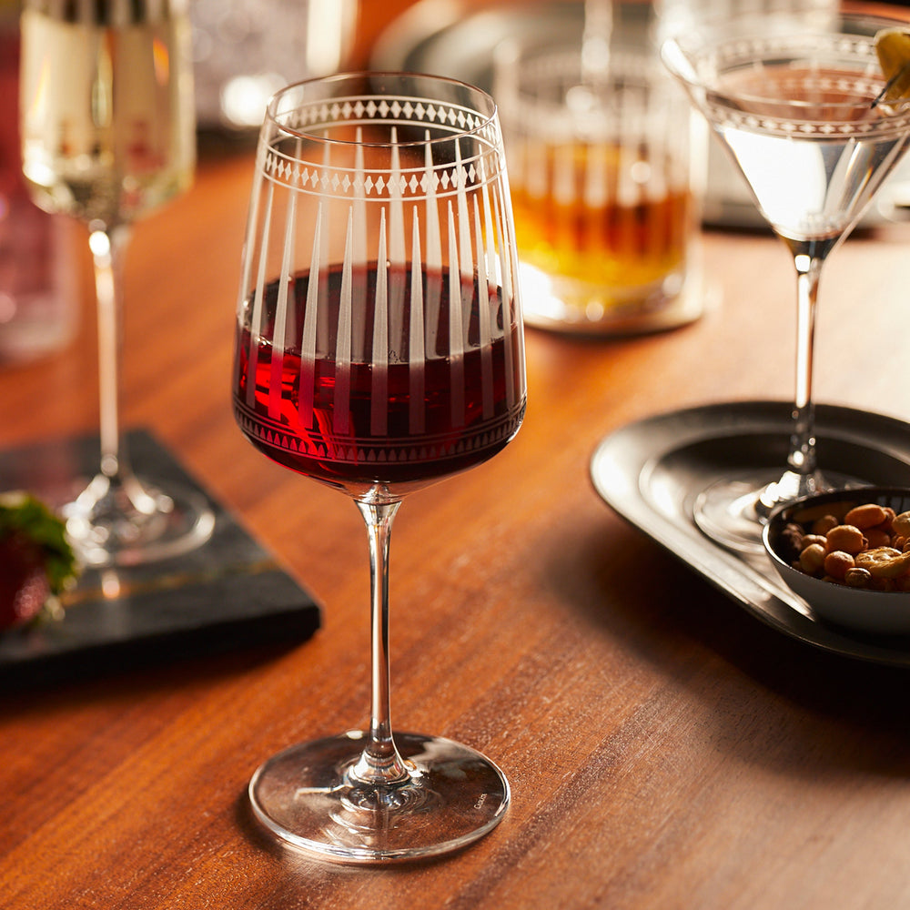A glass of red wine in Caskata's lead-free crystal from the Cocktail Hour Bundle rests on a wooden table, accompanied by assorted drinks and a small dish of mixed nuts.