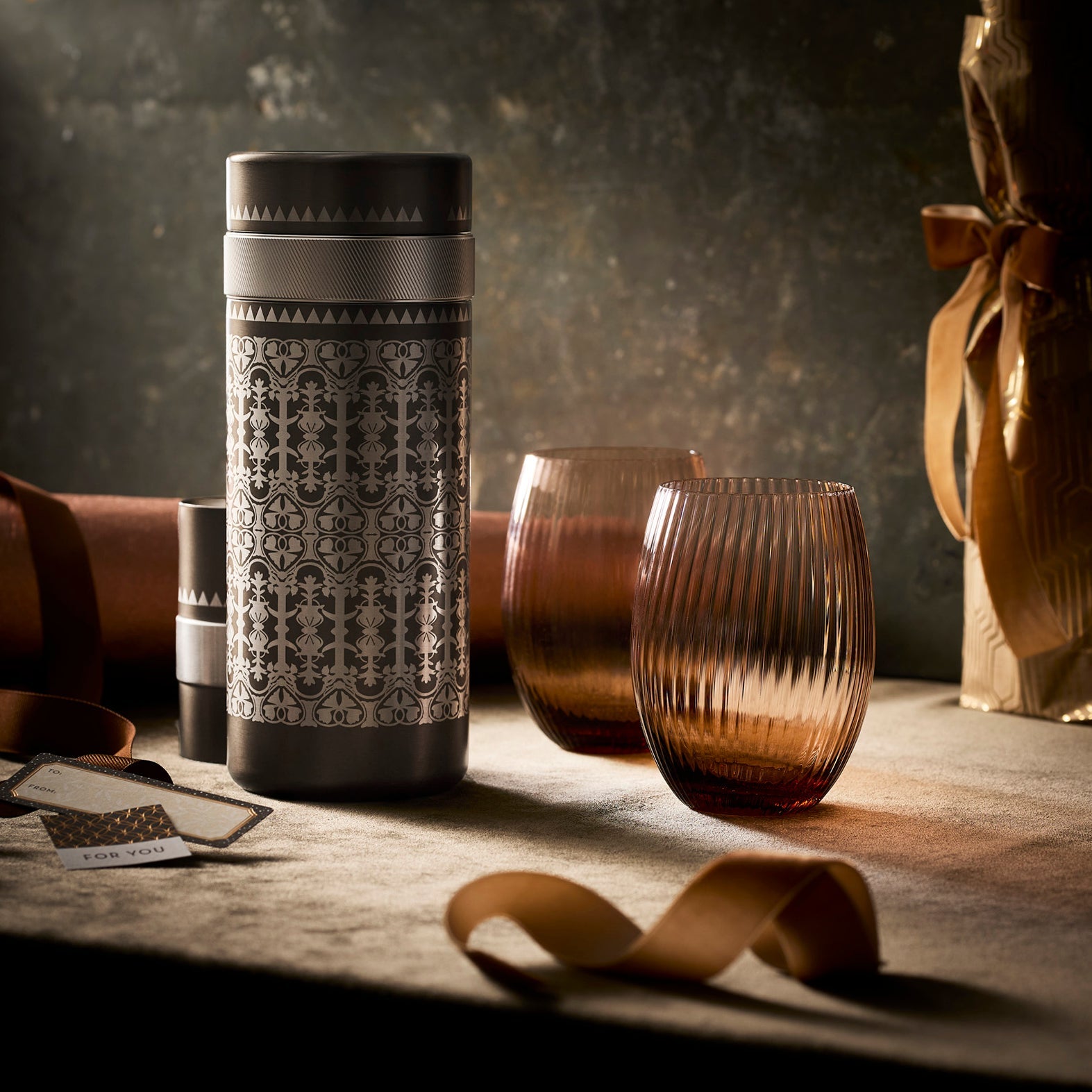 A patterned metal thermos with a screw cap is displayed beside four ribbed amber tumblers on a white background—ideal for serving drinks from your Caskata Golden Hour Bar Bundle.