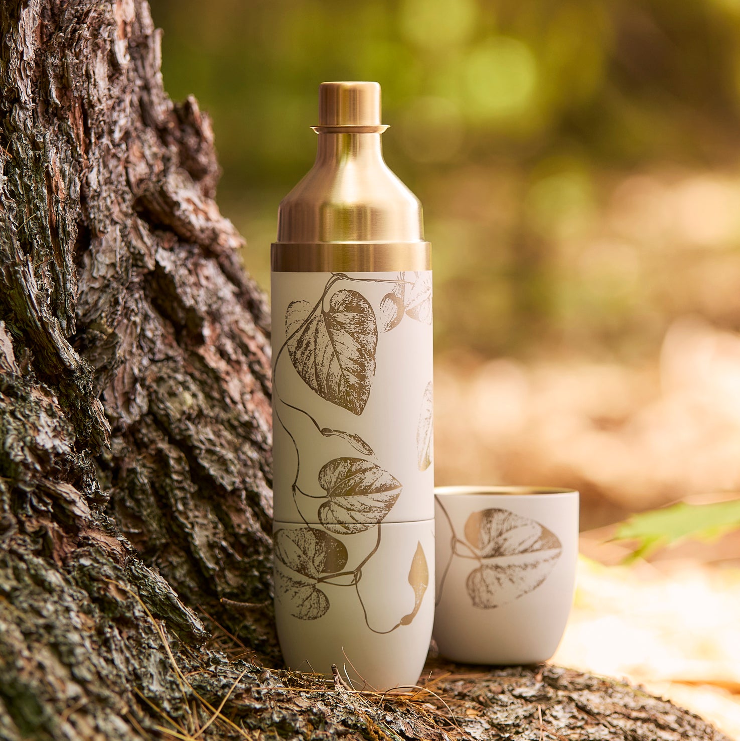 A white and gold leaf-patterned Caskata x High Camp Crema Morning Glory Fern Wine Flask stands upright next to a rectangular black box labeled with the Caskata brand, featuring durable stainless steel construction.
