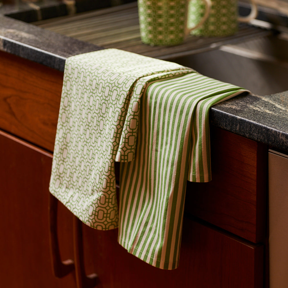 A green and white patterned kitchen towel, part of our Newport Garden Gate & Pinstripe Verde Kitchen Towels, Mixed Set of 2 collection by Caskata, is draped over the edge of a sink with wooden cabinets below. Nearby, a ceramic mug with a similar Newport Garden Gate pattern sits on the counter, making perfect kitchen companions.
