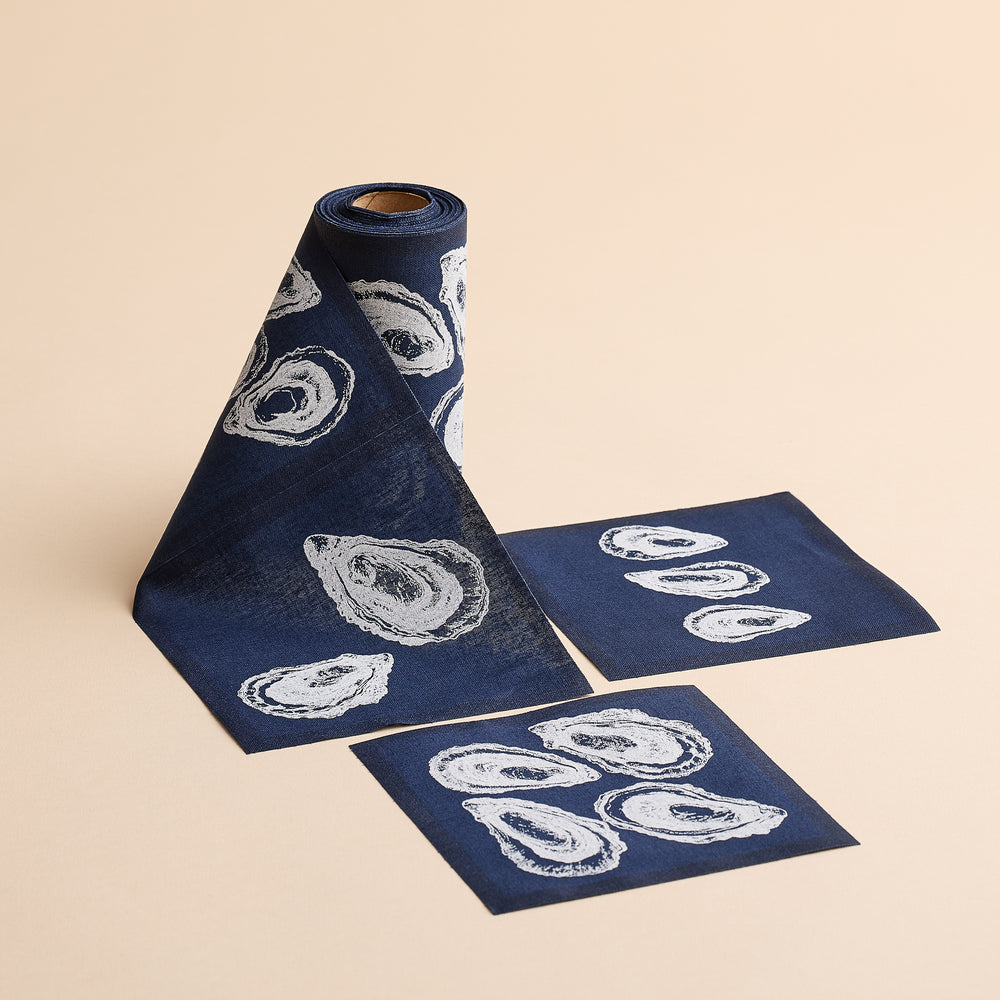 An Oysters Cocktail Napkin Roll by MY DRAP in dark blue with white oyster shell prints, paired with two coordinating square sheets, styled gracefully on a beige surface.