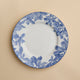 A Caskata Artisanal Home Arbor Rimmed Salad Plate, featuring blue leafy patterns around the white rim, is displayed on a beige background.