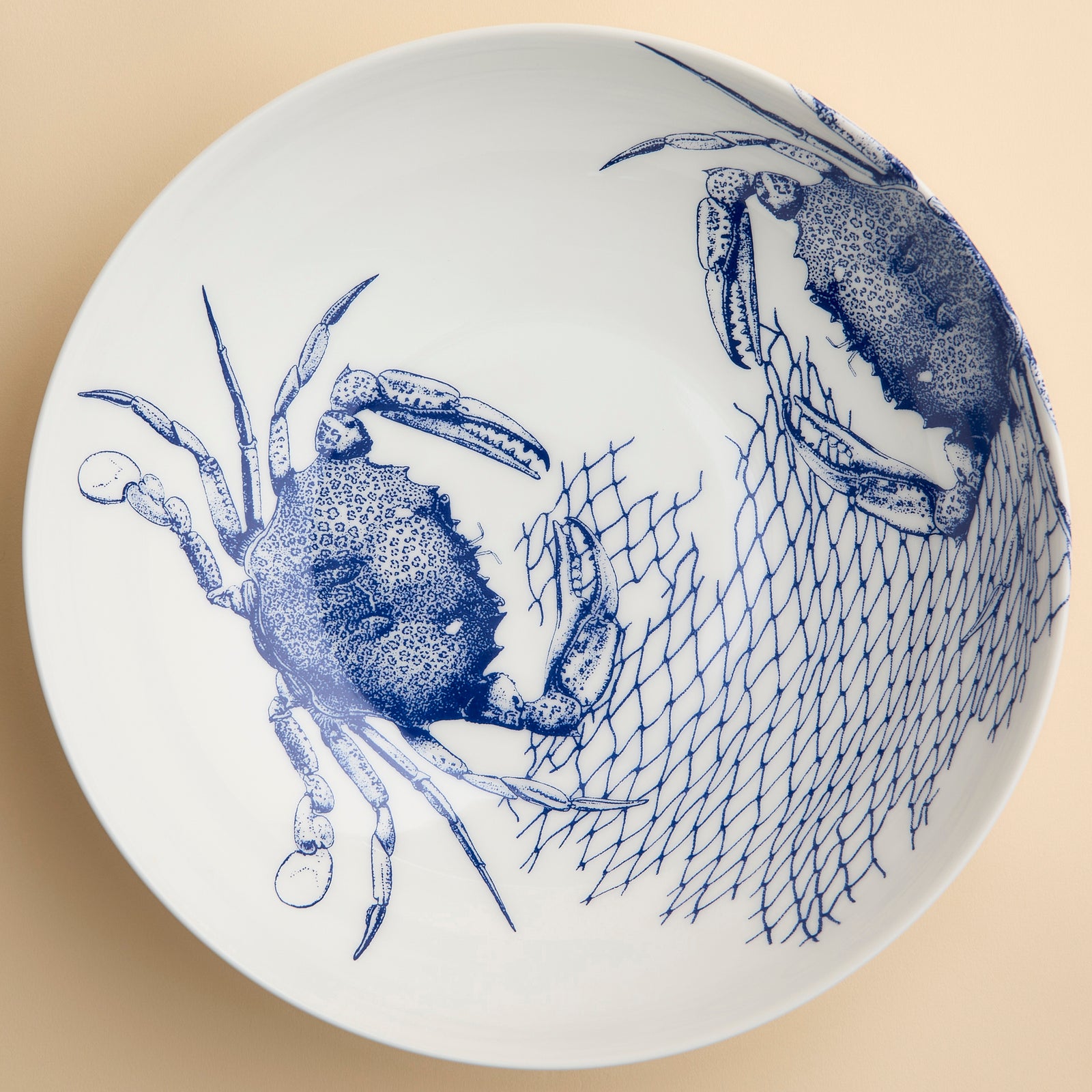 The Crab Wide Serving Bowl by Caskata features white porcelain adorned with blue illustrations of two crabs and a fishing net on the right side, set against a creamy colored background. Reminiscent of coastal charm, this piece is perfect as a decorative serving bowl for any seaside-inspired table setting.