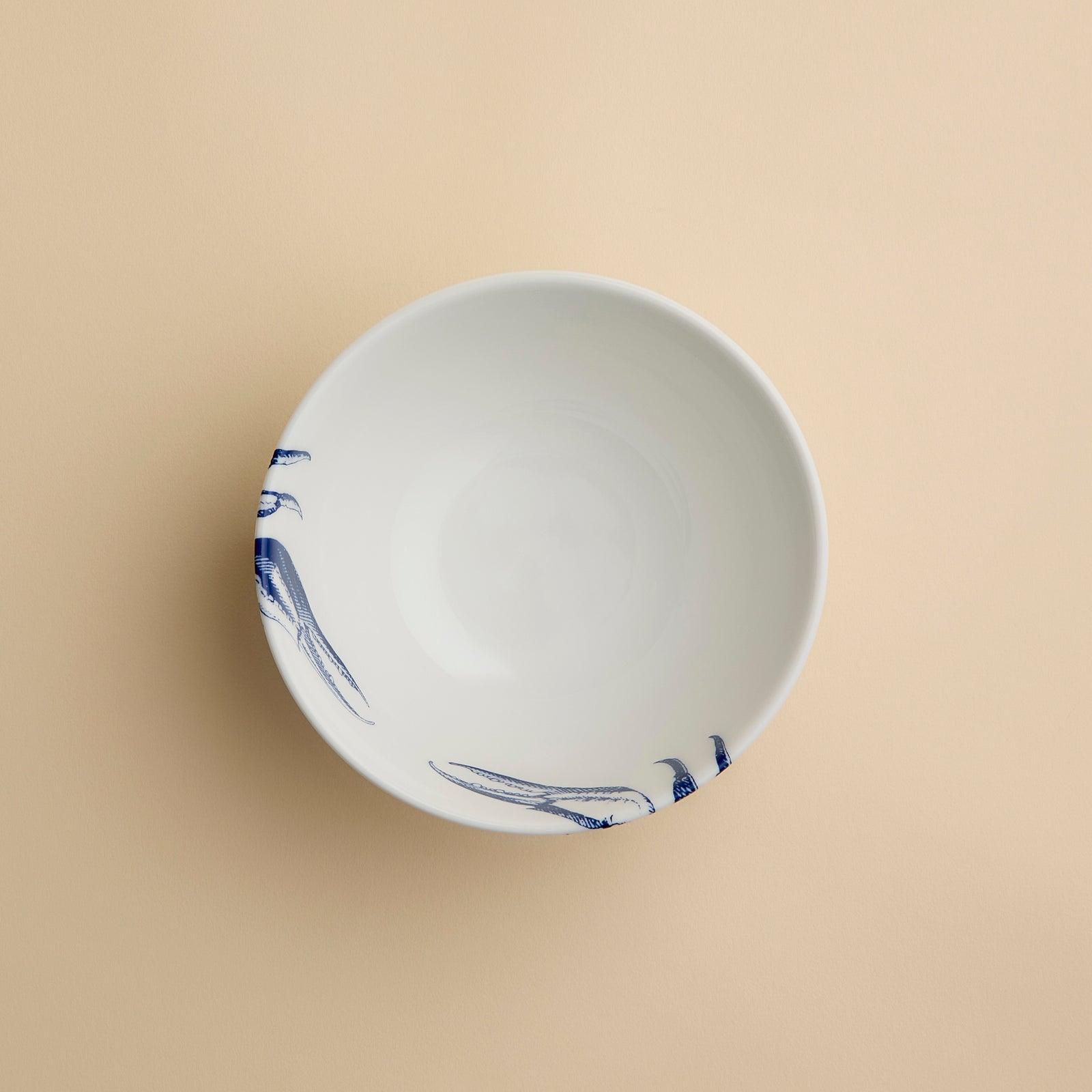 The Caskata Crab Cereal Bowl is a white ceramic bowl featuring a blue illustration of a crab on one side, displayed against a plain beige background.