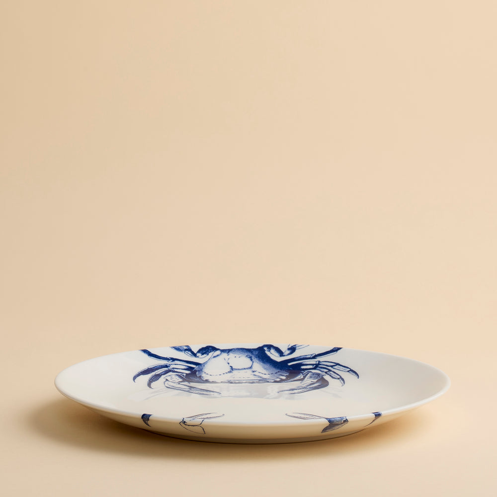 The Crab Coupe Dinner Plate by Caskata Artisanal Home is a white ceramic coastal porcelain plate adorned with a blue illustration of a crab, shown against a plain beige background.
