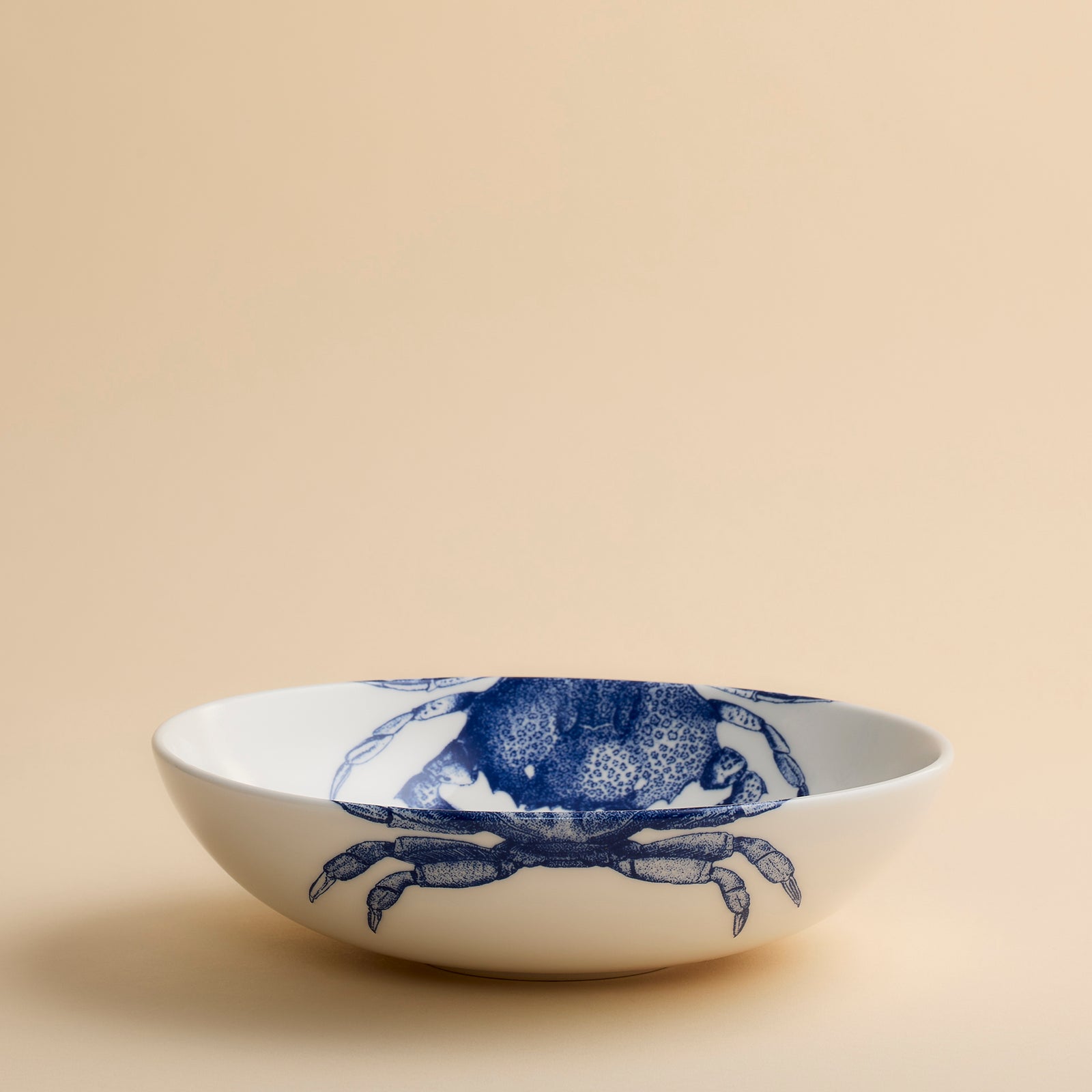 The Caskata Artisanal Home Crab Entrée Bowl is a white ceramic bowl featuring two blue crab illustrations on the rim and is pictured on a beige surface; this porcelain bowl is dishwasher safe for easy cleaning.