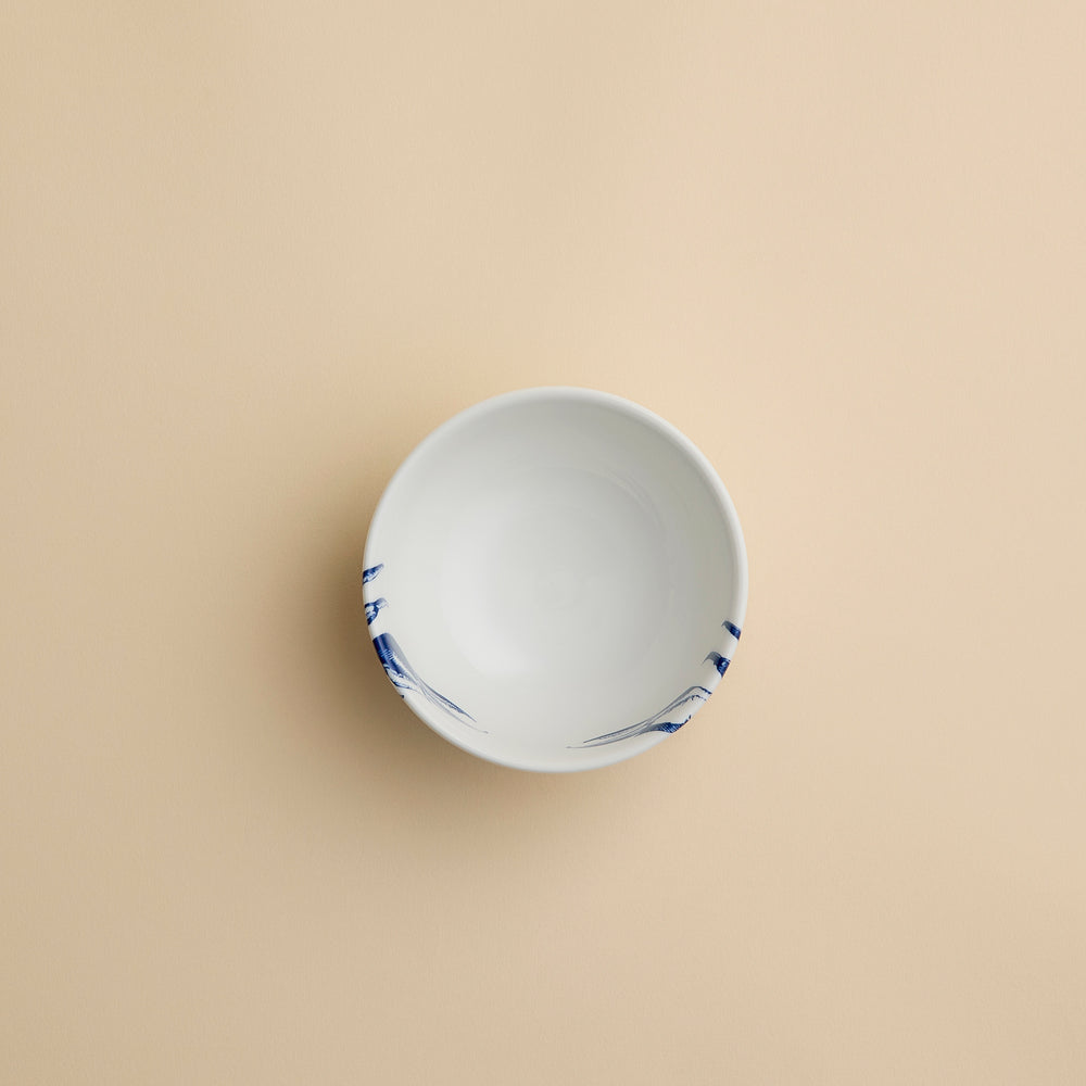 The Caskata Artisanal Home Crab Snack Bowl, a small white porcelain bowl featuring blue brushstroke designs, sits empty on a plain beige background in an overhead view.