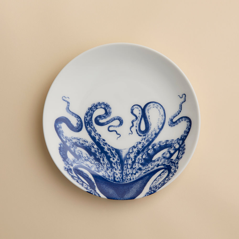 The Lucy Coupe Salad Plate by Caskata Artisanal Home is a white porcelain coupe plate adorned with blue octopus tentacles extending outward on a beige background; this unique salad plate combines stylish design with dishwasher-safe convenience.