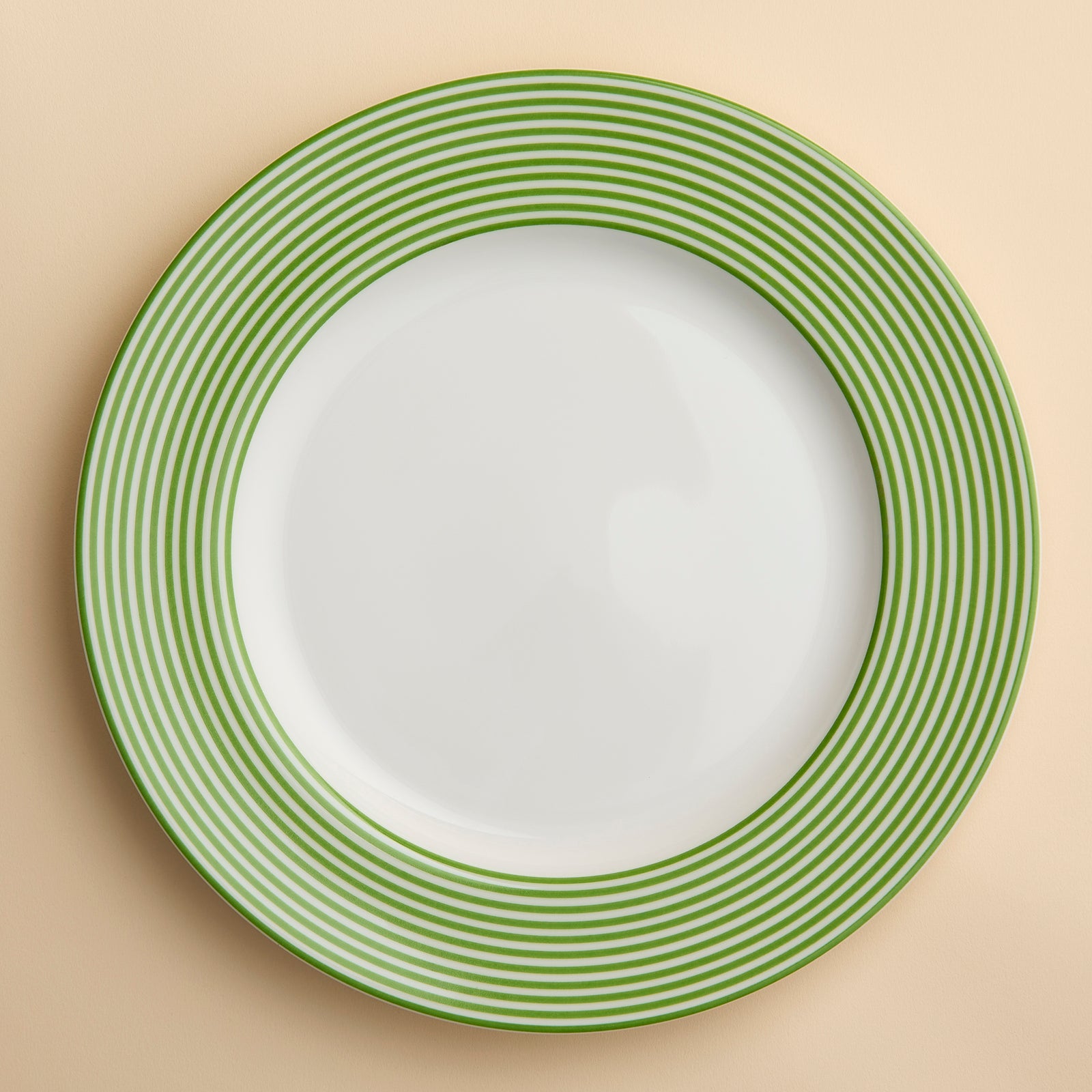 A Newport Stripe Verde Rimmed Dinner Plate featuring the Newport Stripe design with closely spaced green concentric circles around the rim, crafted from high-fired porcelain by Caskata shown against a creamy beige background