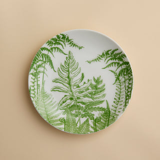 A round white plate adorned with green fern leaf illustrations rests against a beige background—Caskata’s Spring Verde Coupe Salad Plate showcases a charming botanical motif that lends elegance to any table setting. Expertly crafted from premium porcelain, this salad plate blends style with lasting durability.