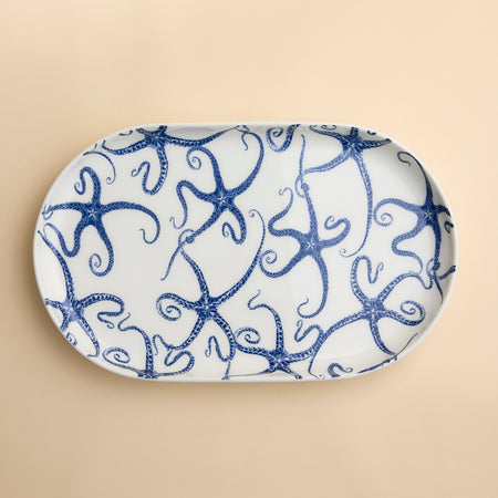 A whimsical oval serving platter with a contemporary form in blue and white porcelain in the best-selling pattern Starfish from Caskata seen overhead against a beige background