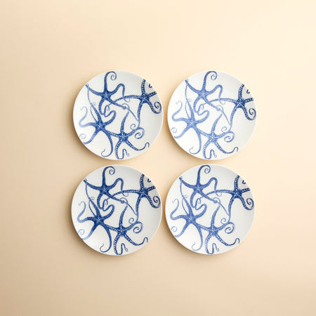 Whimsical Starfish dance across the faces of this set of 4 small appetizer plates from Caskata in creamy blue and white porcelain against a beige background.