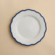 A white porcelain salad plate with a wavy, dark blue rim—the Stella Scalloped Blue Salad Plate by Caskata—is centered on a light beige background. This plate combines elegance and practicality, as it is both microwave and dishwasher safe.