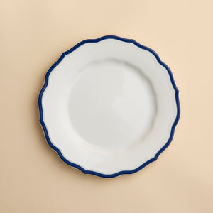 A white porcelain salad plate with a wavy, dark blue rim—the Stella Scalloped Blue Salad Plate by Caskata—is centered on a light beige background. This plate combines elegance and practicality, as it is both microwave and dishwasher safe.