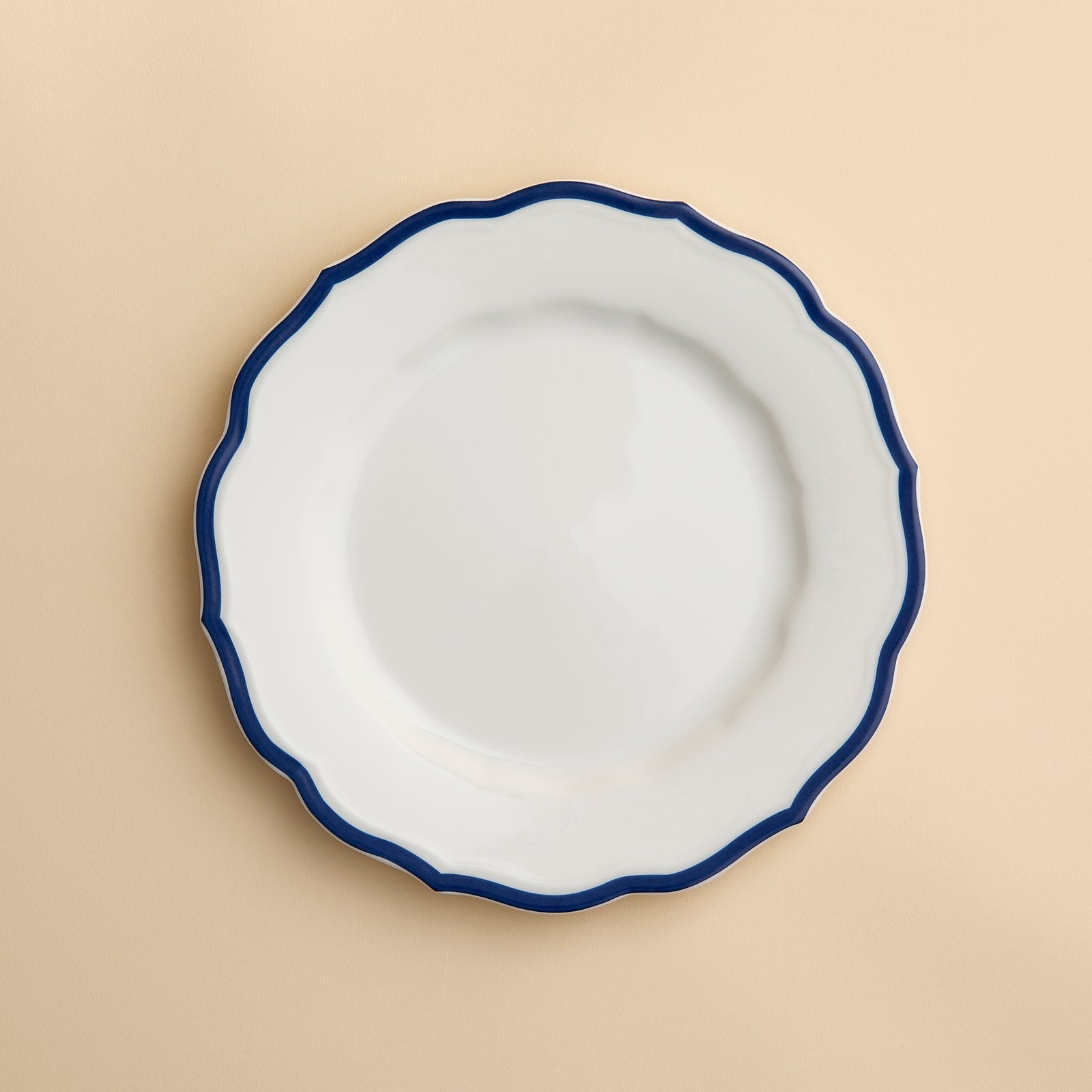 A white porcelain salad plate with a wavy, dark blue rim—the Stella Scalloped Blue Salad Plate by Caskata—is centered on a light beige background. This plate combines elegance and practicality, as it is both microwave and dishwasher safe.