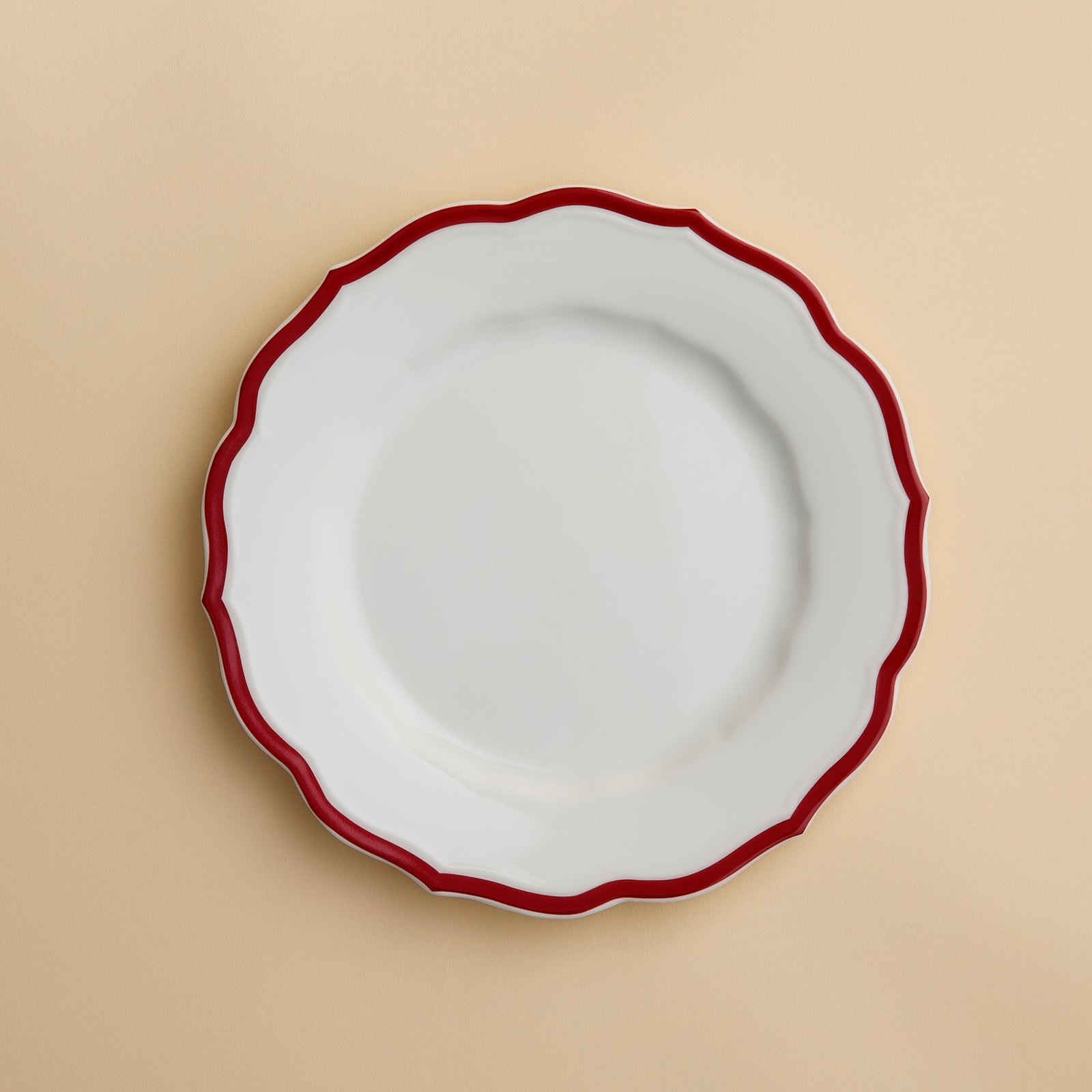 The Stella Scalloped Crimson Salad Plate by Caskata, featuring a white ceramic surface with a wavy crimson rim inspired by vintage dinnerware, is displayed on a light beige background.