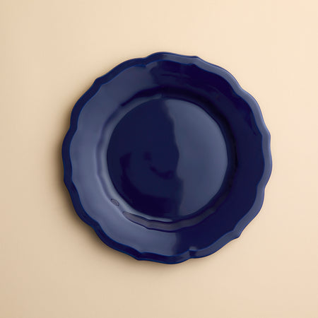 A  dark blue Caskata Stella Scalloped Solid Blue Salad Plate with a scalloped edge in porcelain on a creamy beige background.