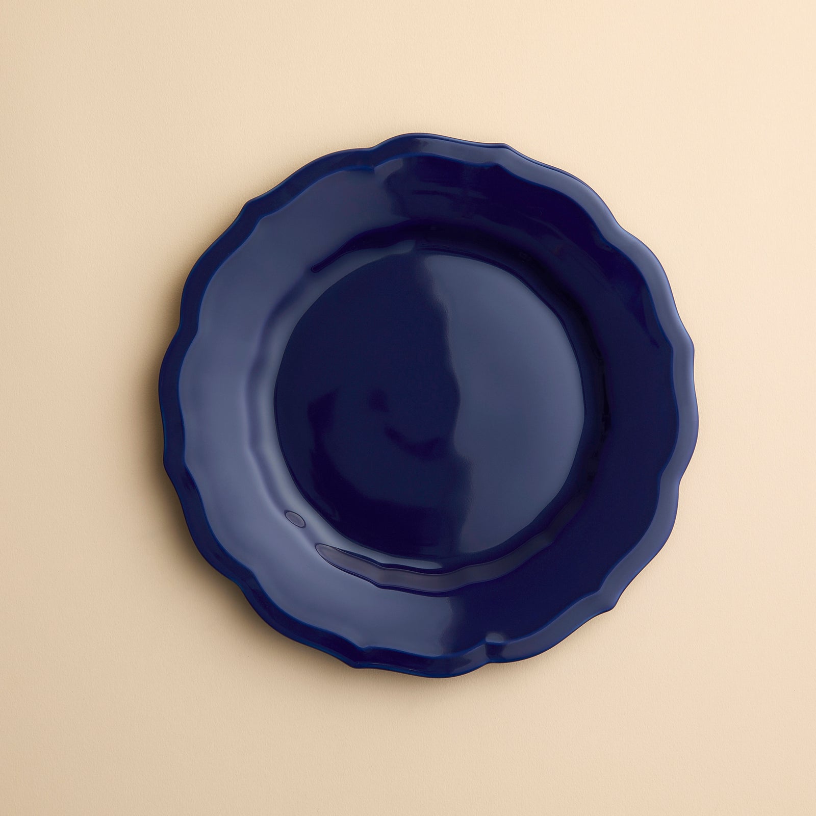 A  dark blue Caskata Stella Scalloped Solid Blue Salad Plate with a scalloped edge in porcelain on a creamy beige background.
