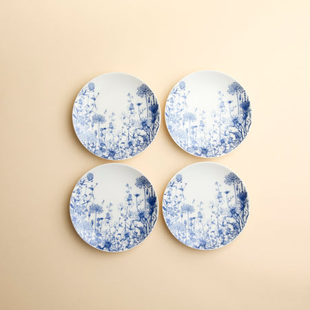 Set of 4 Summer Blues Small Plates by Caskata, crafted from high-fired porcelain with blue floral designs, are arranged against a creamy beige background