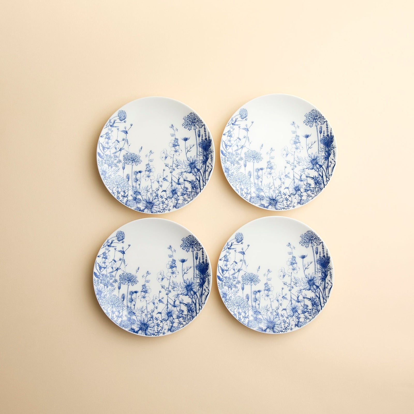 Set of 4 Summer Blues Small Plates by Caskata, crafted from high-fired porcelain with blue floral designs, are arranged against a creamy beige background