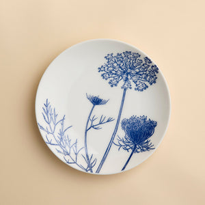 White ceramic plate adorned with blue botanical illustrations of flower stems and leaves, set on a beige background. The Summer Blues Coupe Salad Plate by Caskata Artisanal Home brings a sophisticated botanical touch to your porcelain dinnerware collection.