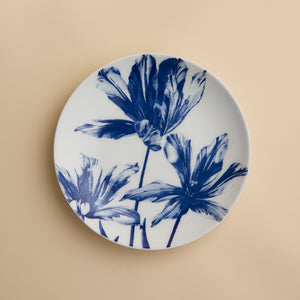 The Caskata Tulip Coupe Salad Plate is a white ceramic plate adorned with a blue floral design featuring three large flowers, beautifully displayed against a plain beige background.