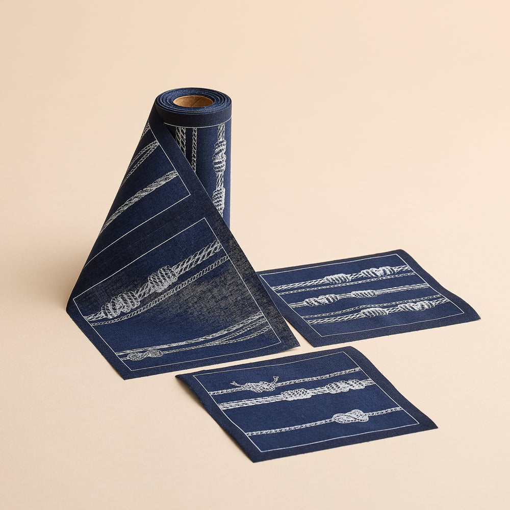 A partially unrolled MY DRAP Sailors Knots Cocktail Napkin Roll—navy blue with white rope designs—rests on a beige background, accompanied by two matching square napkins, ideal as reusable party or cotton cocktail napkins.
