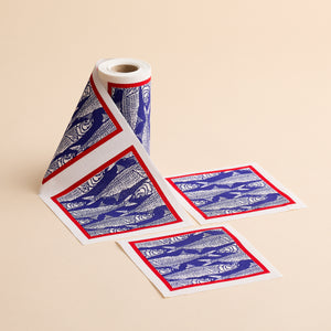 A roll of patterned paper from MY DRAP, the Sardines Cocktail Napkin Roll, showcases a charming block print fish design with blue fish illustrations and red borders. Two individual square sheets—resembling cotton cocktail napkins—have been torn off and are placed beside the roll on a beige surface.