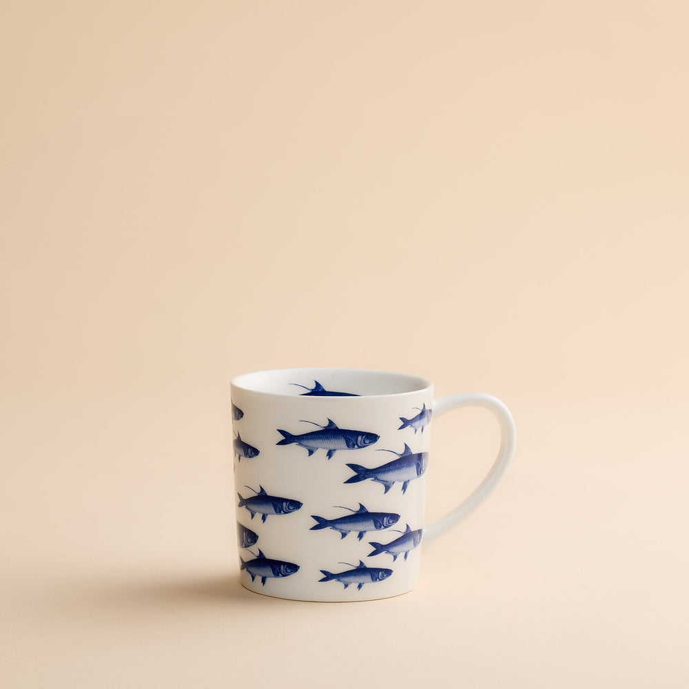 The School of Fish Mug by Caskata Artisanal Home features blue fish illustrations on a white ceramic surface and is displayed against a plain beige background. Made from high-fired porcelain, this coastal mug ensures both durability and enduring charm.