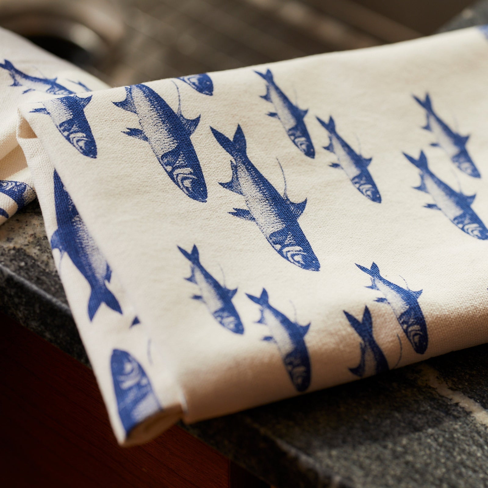 The Caskata Fishing Fun Bundle, including a School of Fish porcelain platter and matching kitchen towel, rests on a rustic wooden table beside gift wrap, ribbon, and a decorative branch.