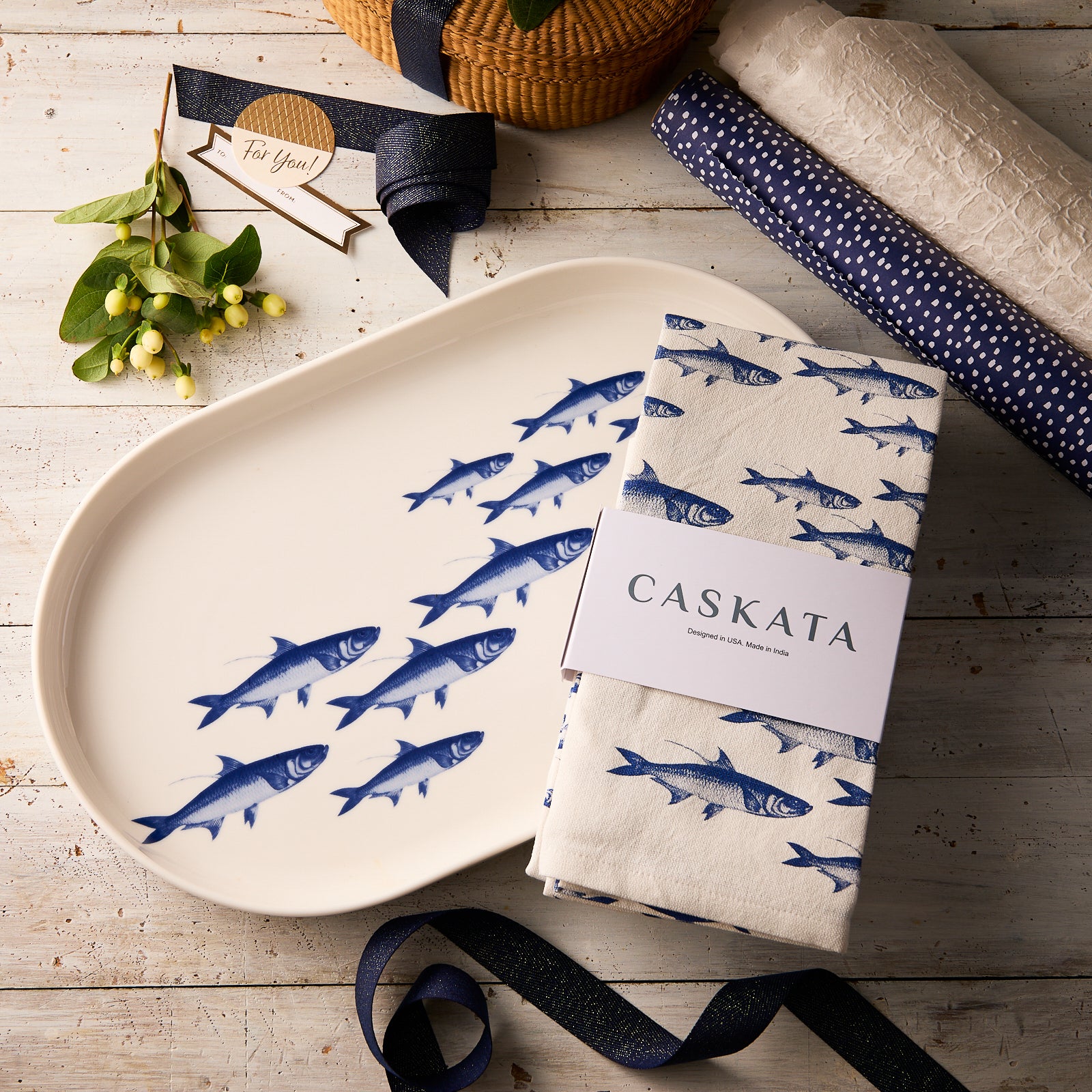 The Caskata Fishing Fun Bundle, including a School of Fish porcelain platter and matching kitchen towel, rests on a rustic wooden table beside gift wrap, ribbon, and a decorative branch.