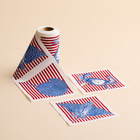The Seaside Stripes Cocktail Napkin Roll by MY DRAP features a roll of patterned napkins adorned with red stripes alongside blue eagle, heart, and deer motifs, reminiscent of coastal charm; two individual sheets are displayed flat on a beige background.