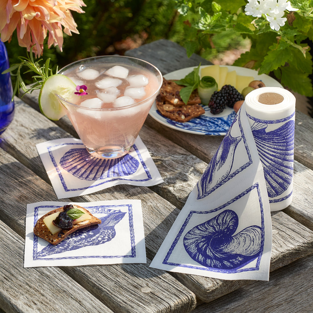 A drink with ice, a cracker topped with cheese and fruit, and a MY DRAP Shells Cocktail Napkin Roll—all made from 100% cotton napkins—are displayed as true party essentials on a weathered wooden outdoor table.
