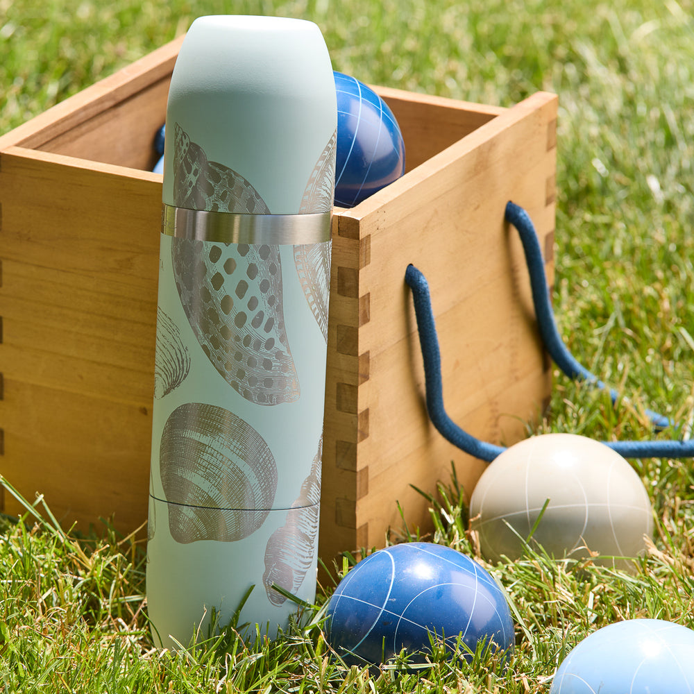 The Caskata x High Camp Light Blue Shells Wine Flask by Caskata, featuring seashell designs, stands on grass beside a wooden box and blue and white bocce balls.