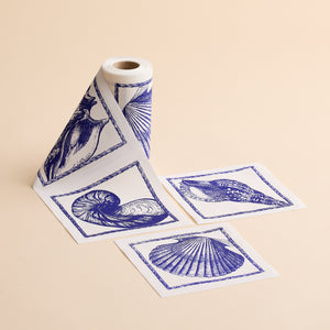 A roll of MY DRAP Shells Cocktail Napkin Roll, decorated with blue seashell and marine designs, is displayed with three square napkins cut and arranged beside it on a beige background—these napkins are a must-have party essential for any celebration.
