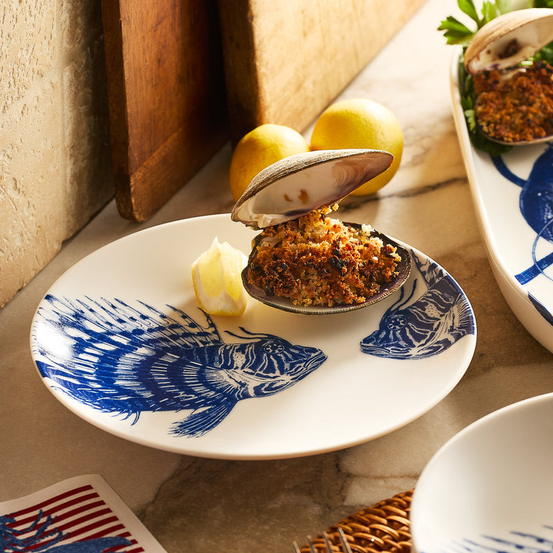 A baked stuffed clam with breadcrumbs and a lemon wedge is served on a Caskata Spiky Fish Coupe Salad Plate, featuring blue fish artwork. Two whole lemons appear in the background.