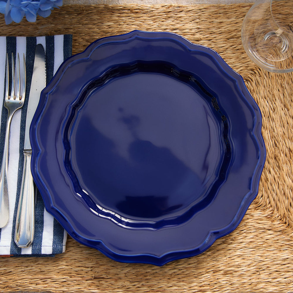 A blue Stella Scalloped Solid Blue Dinner Plate set on a woven placemat next to a striped napkin and silver cutlery.