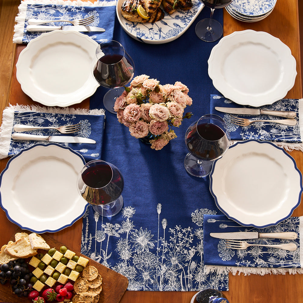 A dining table set for four with white plates, red wine glasses, blue floral placemats, cottage-core linens, a flower centerpiece, assorted appetizers, and Caskata Summer Blues Dinner Napkins that are machine washable.