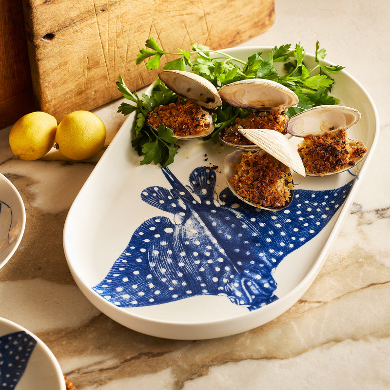 The Caskata Ray the Stingray Large Coupe Oval Platter holds baked stuffed clams, fresh parsley, and lemon halves, displayed on a marble countertop.