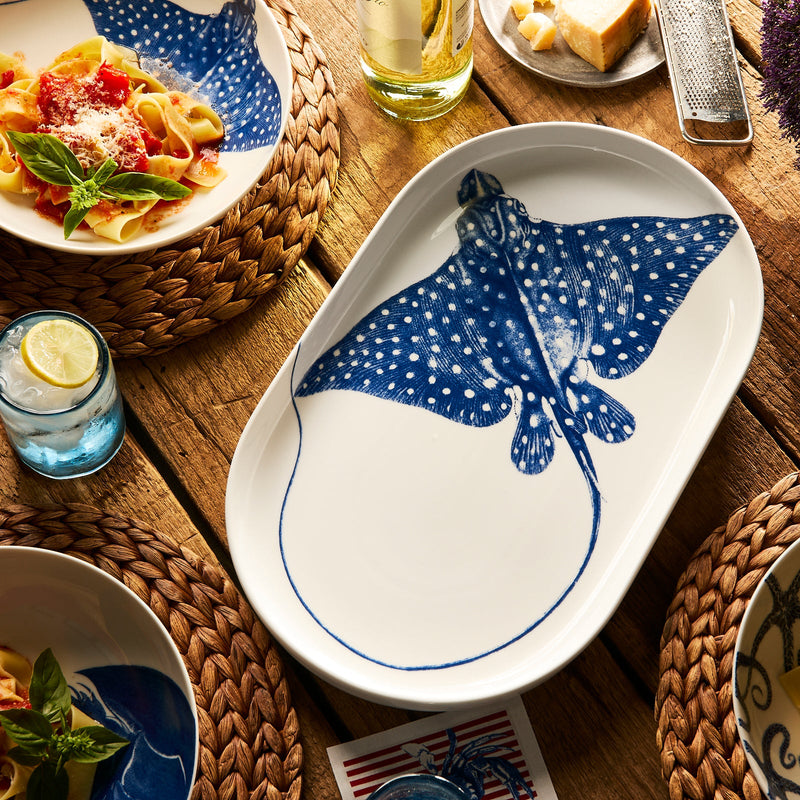 The Caskata "Ray the Stingray" Large Coupe Oval Platter, featuring a blue stingray design, adds a coastal touch to your table and is shown on a wooden table with pasta, drinks, and woven placemats.