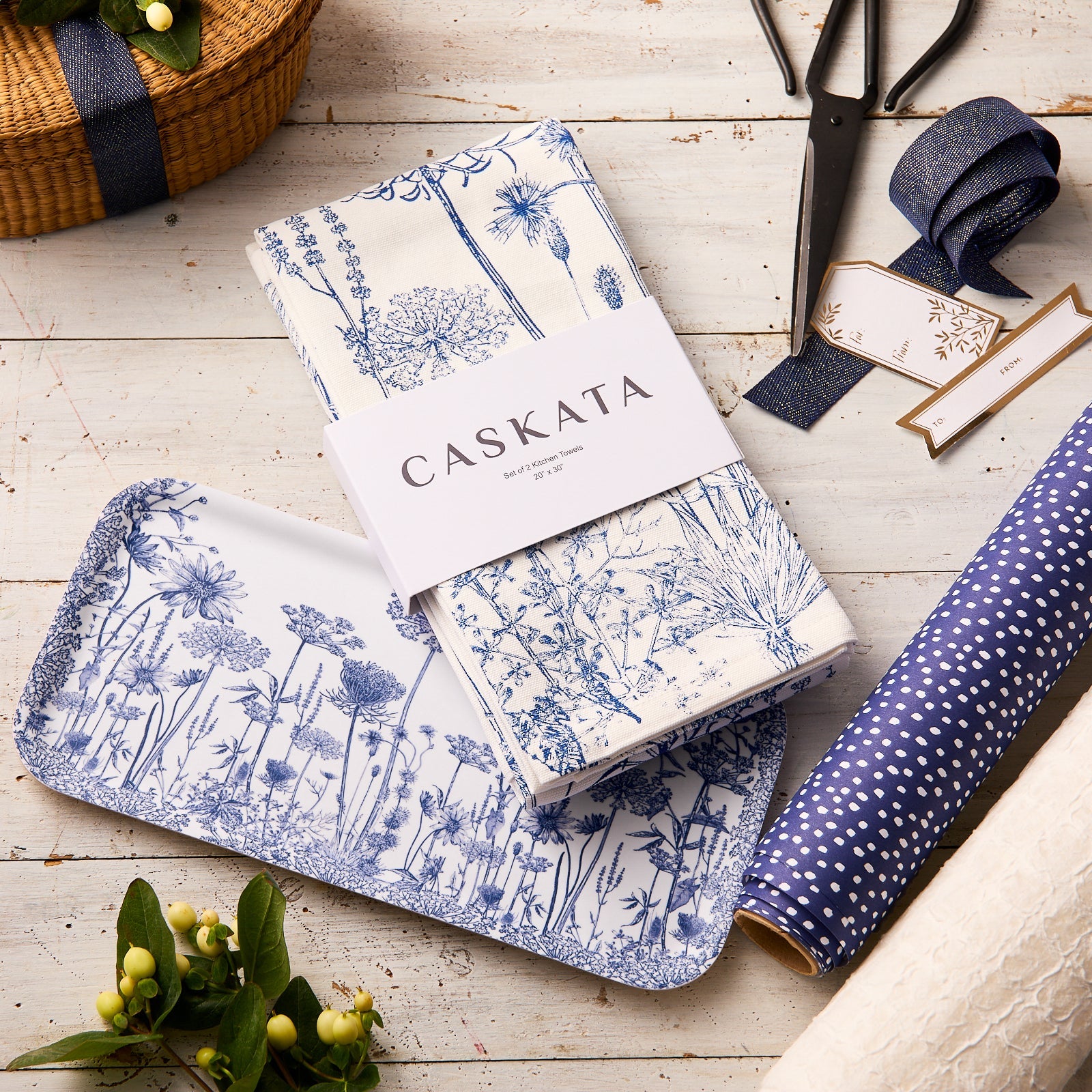 The Caskata Blooming Blues Bundle, featuring a wildflower serving tray and matching blue botanical cotton kitchen towels, is styled with ribbon, scissors, and gift wrap on a rustic white table for a charming cottagecore look.