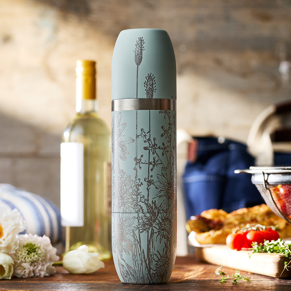 A Caskata x High Camp Light Blue Summer Wine Flask from Caskata stands on a wooden table, accompanied by a bottle of white wine, food, flowers, and kitchen items softly blurred in the background.