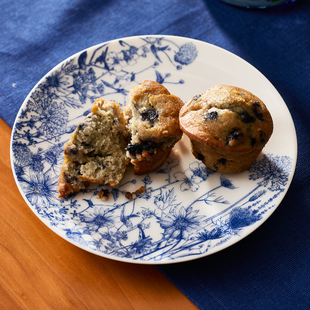 Two blueberry muffins, one whole and one split, rest on a Caskata Summer Blues Small Plate atop a wooden table with a blue cloth.