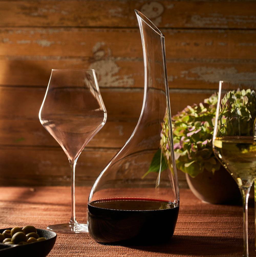 Caskata Terrace Red Wine Glasses (Set of 4) are arranged on a table with a glass wine decanter, a bowl of olives, and flowers, all set against a wooden wall.
