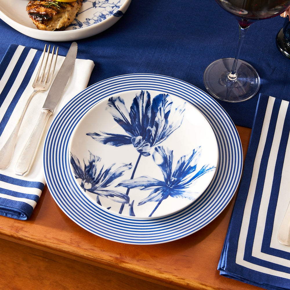 A table setting featuring Caskata's Tulip Coupe Salad Plate, a blue floral plate, striped napkins, cutlery, a glass of red wine, and grilled food.